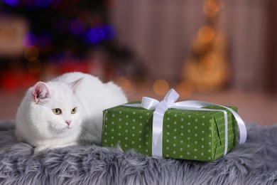 Photo of Christmas atmosphere. Cute cat lying near gift box on fur rug against blurred lights. Space for text