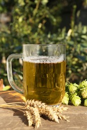 Photo of Mug with beer, fresh hops and ears of wheat on wooden table outdoors