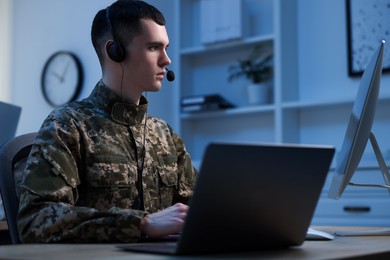 Photo of Military service. Young soldier in headphones working at wooden table in office at night