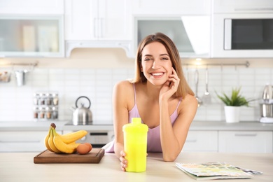 Photo of Young woman holding bottle of protein shake at table with ingredients in kitchen