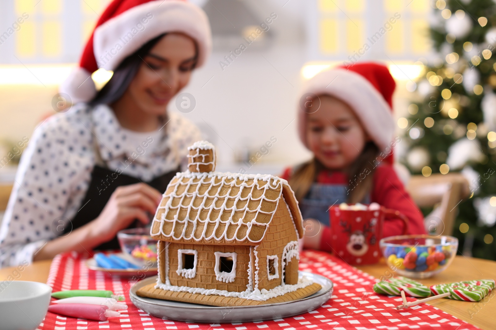 Photo of Mother and daughter in Santa hats cooking together, focus on gingerbread house
