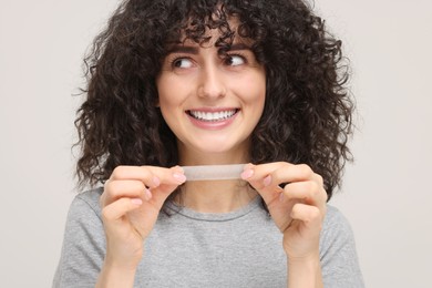 Young woman holding teeth whitening strip on light grey background