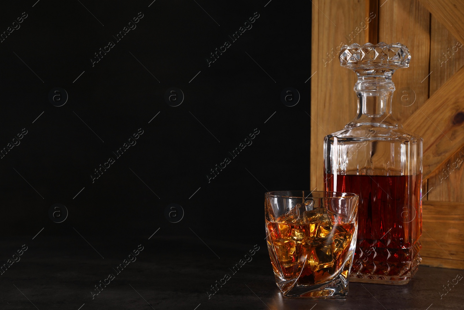 Photo of Whiskey in glass and bottle near wooden crate on dark table against black background. Space for text