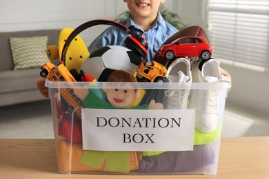 Photo of Little boy holding donation box with goods and toys at home, closeup