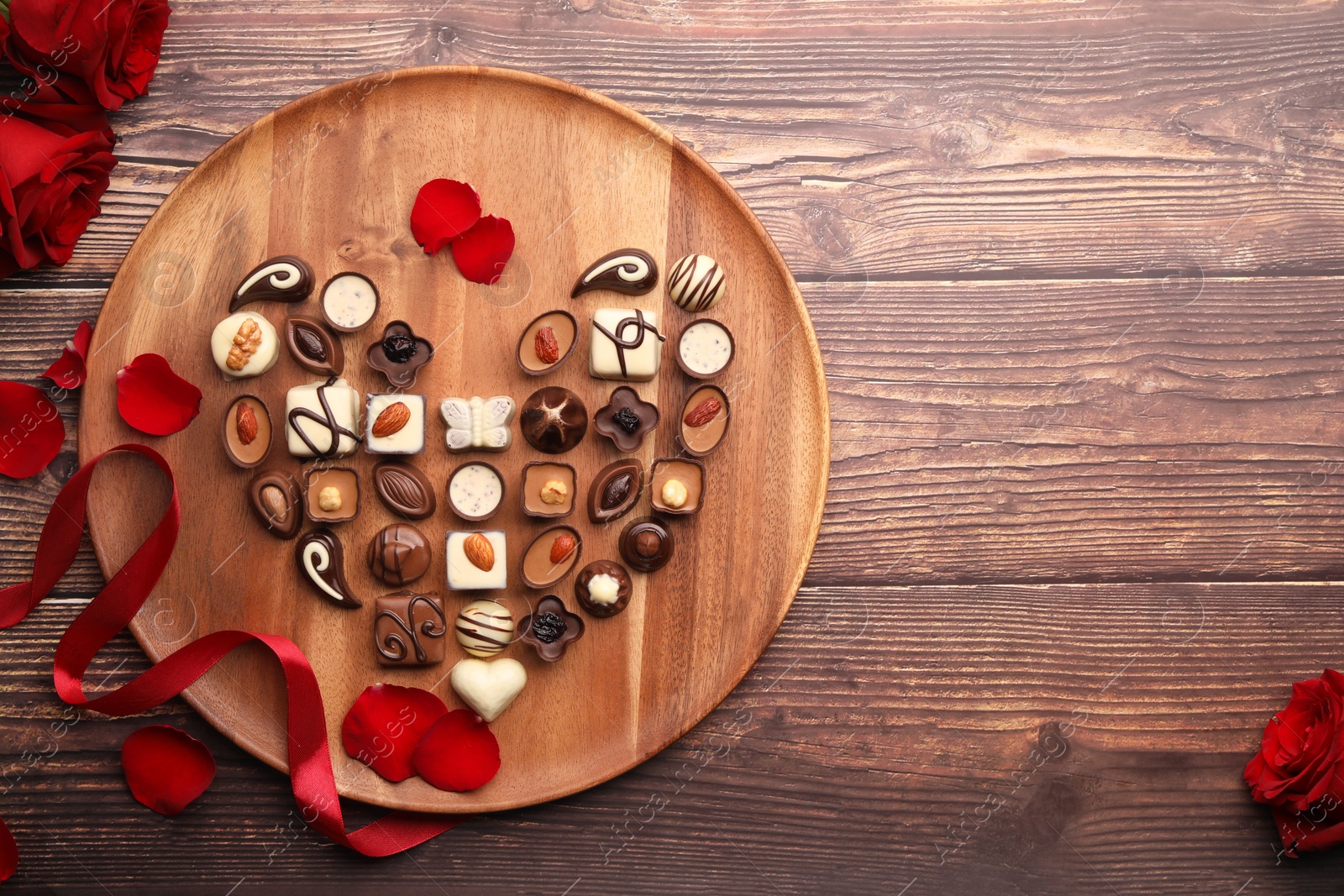 Photo of Heart made with delicious chocolate candies and rose petals on wooden table, top view. Space for text