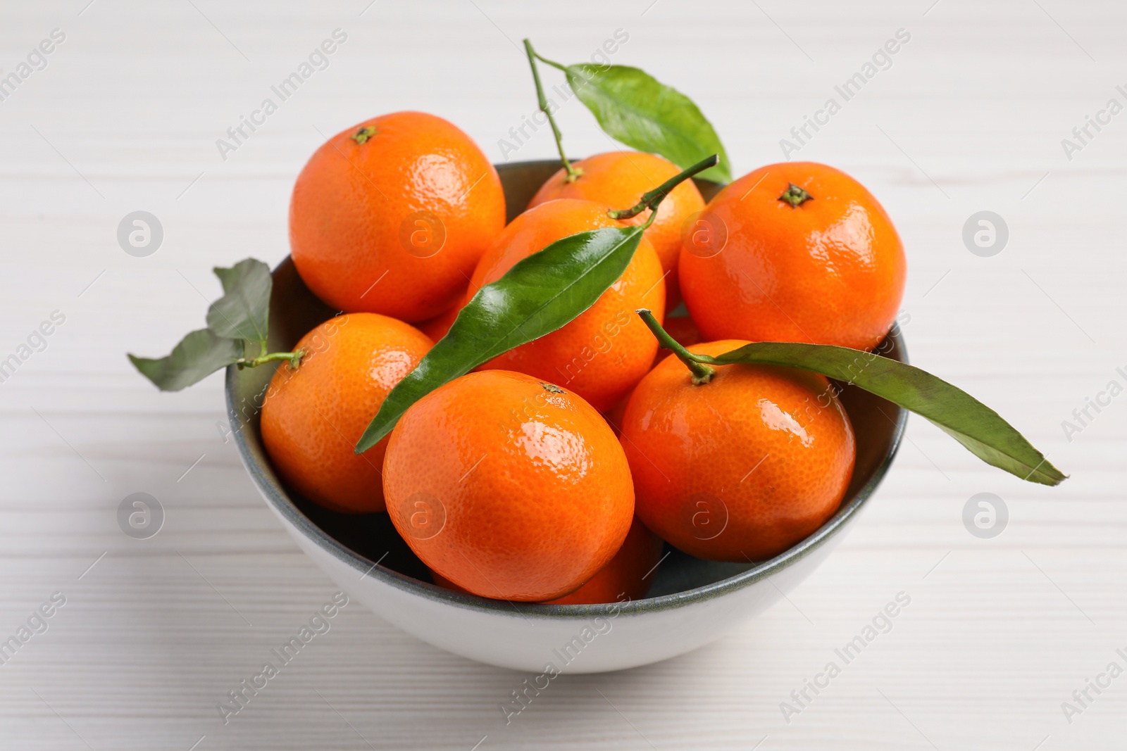 Photo of Delicious tangerines with green leaves in bowl on white wooden table, closeup