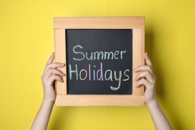 Schoolgirl holding blackboard with text SUMMER HOLIDAYS on yellow background, closeup