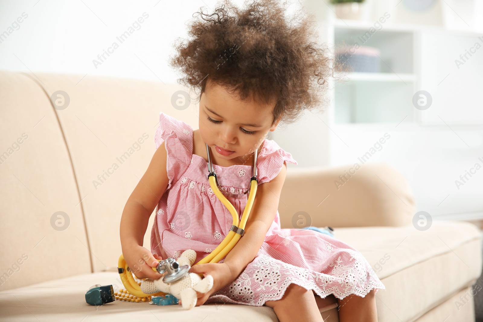 Photo of Cute African American child imagining herself as doctor while playing with stethoscope and toy on couch at home