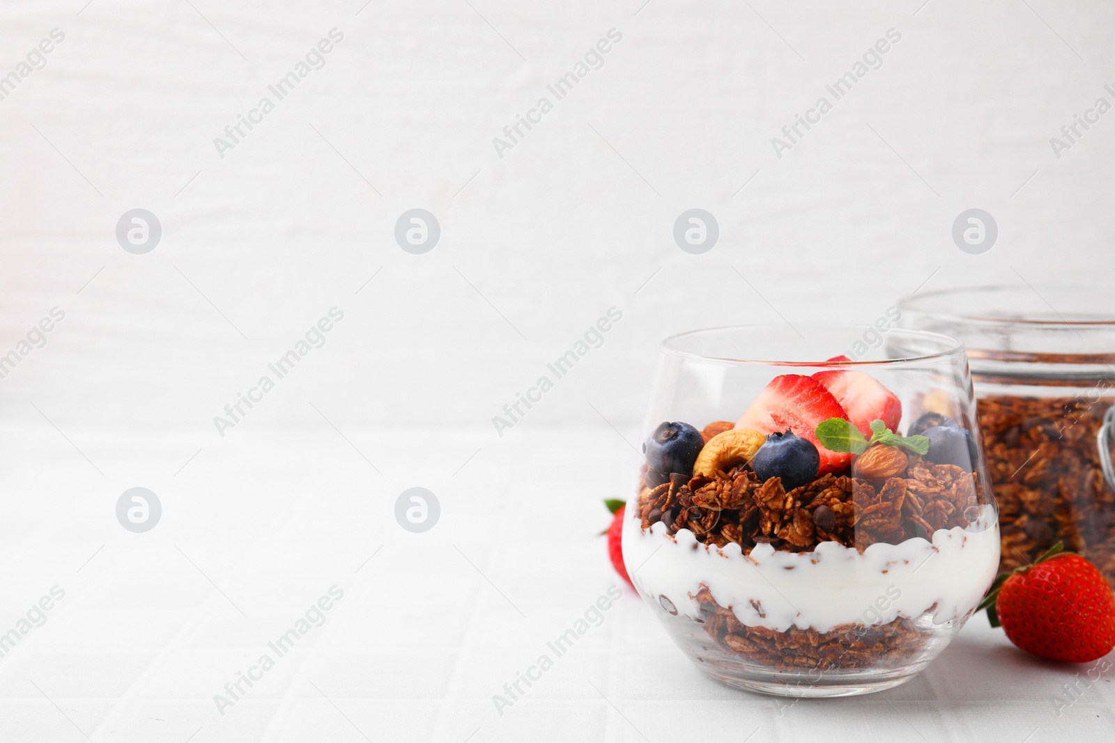 Photo of Tasty granola with berries, nuts and yogurt in glass on white tiled table, space for text