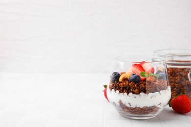 Photo of Tasty granola with berries, nuts and yogurt in glass on white tiled table, space for text