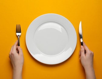 Woman with empty plate and cutlery on orange background, top view