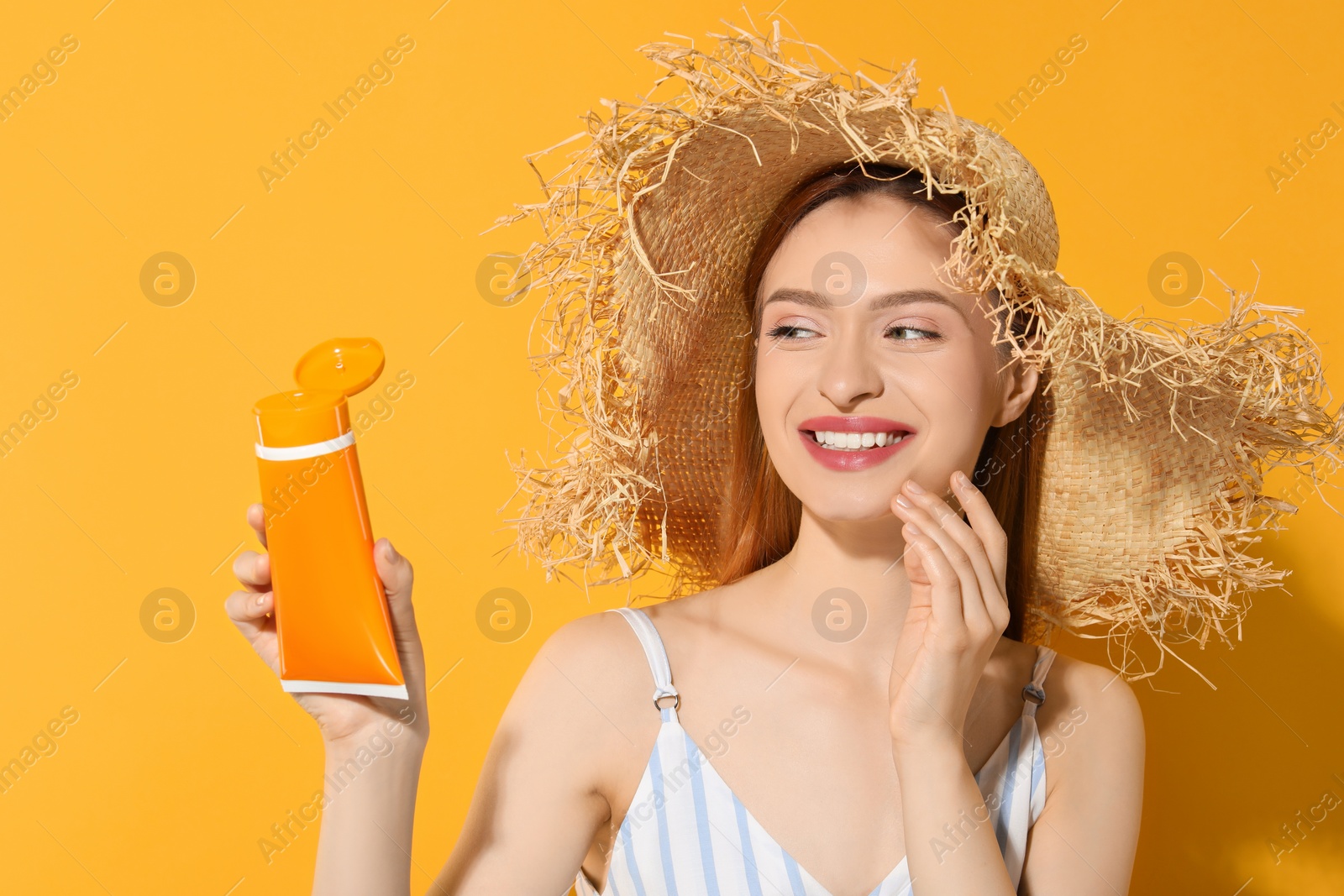 Photo of Beautiful young woman in straw hat holding sun protection cream on orange background