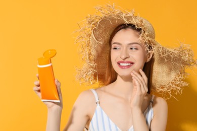 Beautiful young woman in straw hat holding sun protection cream on orange background