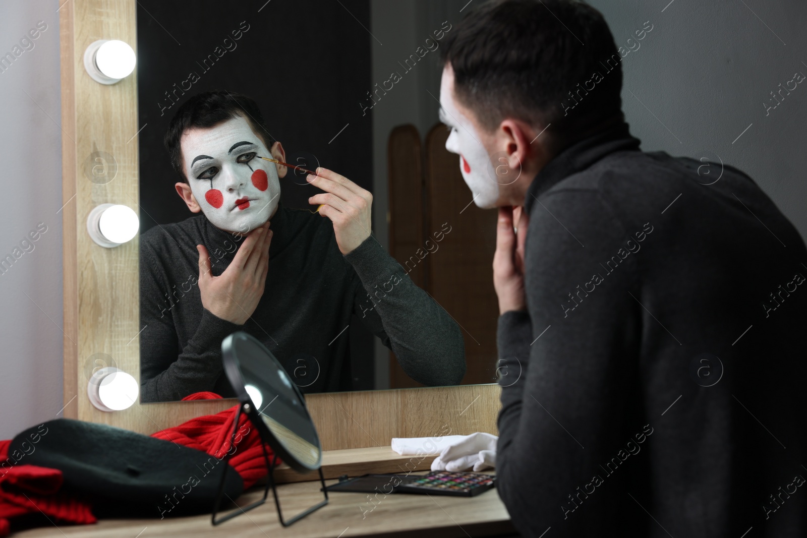 Photo of Young man applying mime makeup near mirror in dressing room