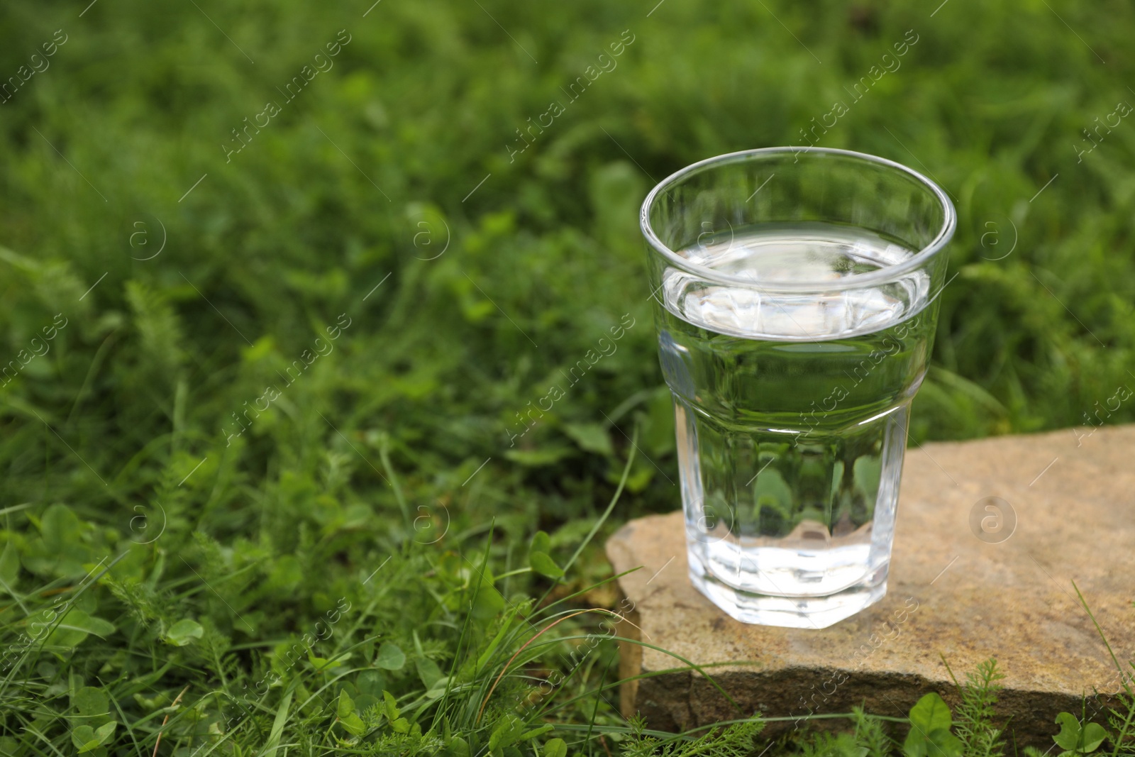 Photo of Glass of fresh water on stone in green grass outdoors. Space for text