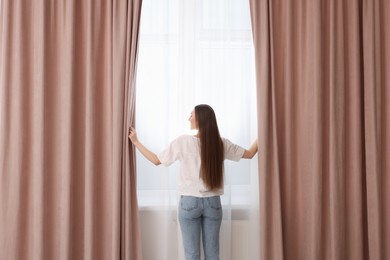 Photo of Woman opening stylish curtains at home, back view