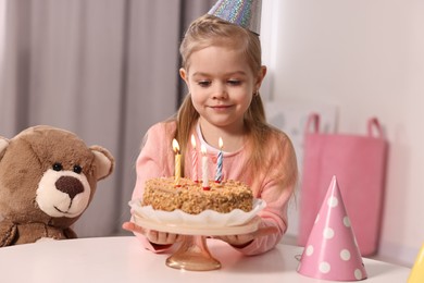 Photo of Cute girl in party hat with birthday cake at table indoors