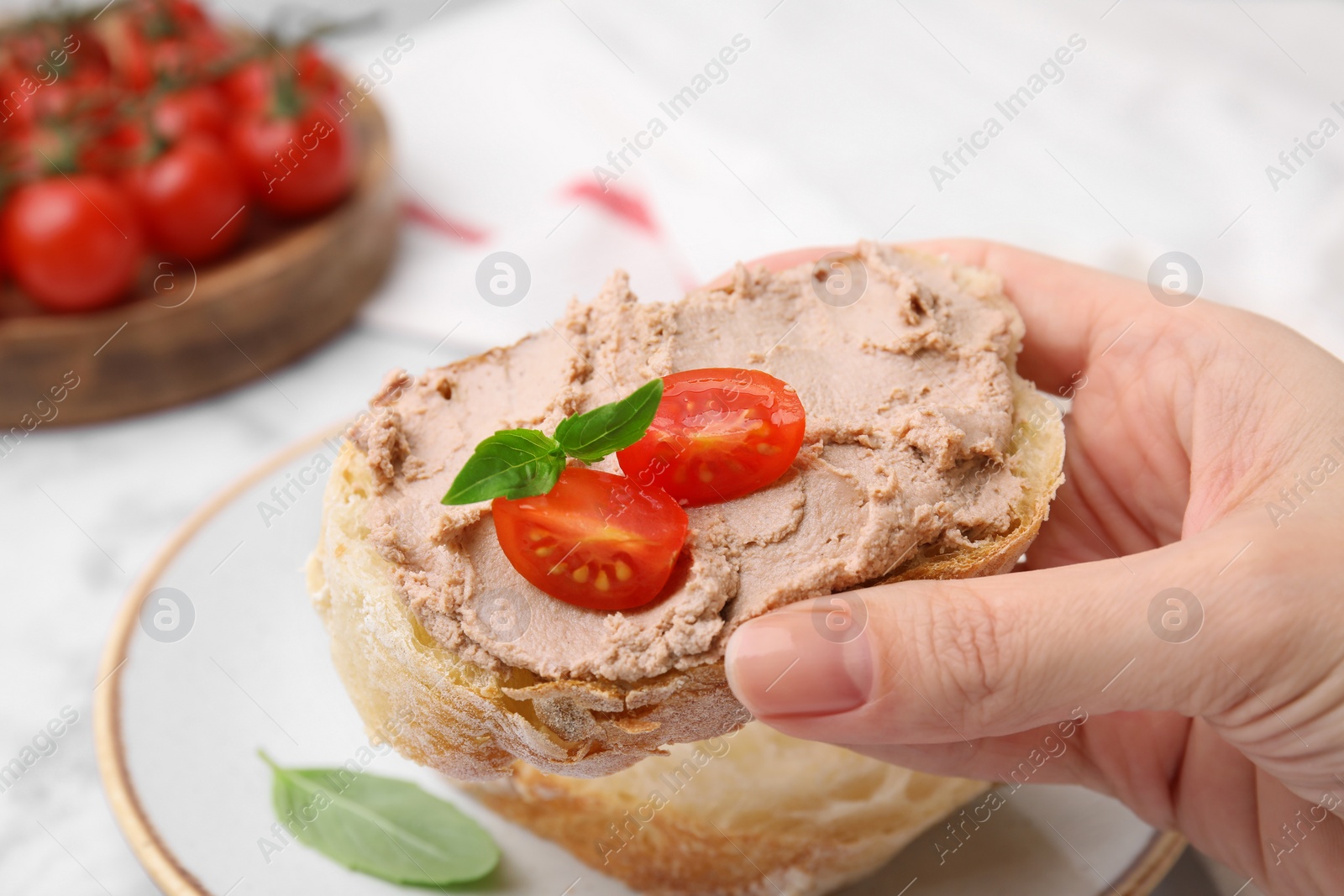 Photo of Woman holding delicious liverwurst sandwich with tomatoes and basil at table, closeup