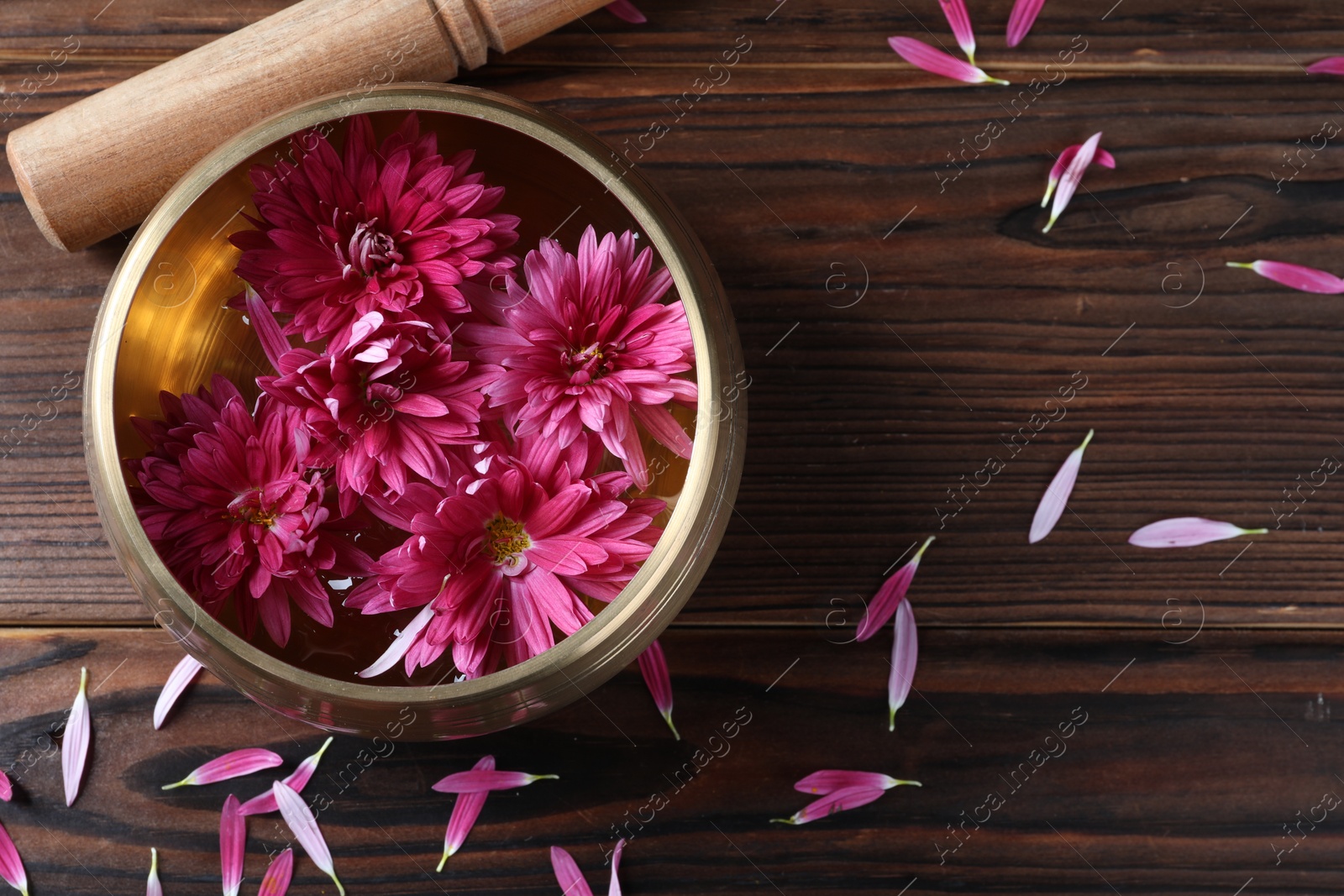 Photo of Tibetan singing bowl with water, beautiful flowers and mallet on wooden table, top view. Space for text