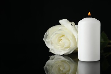 White rose and burning candle on black mirror surface in darkness, closeup with space for text. Funeral symbols