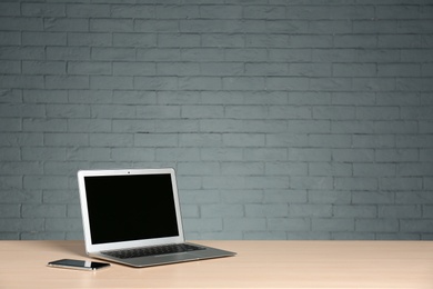 Modern laptop with blank screen and mobile phone on table against brick wall