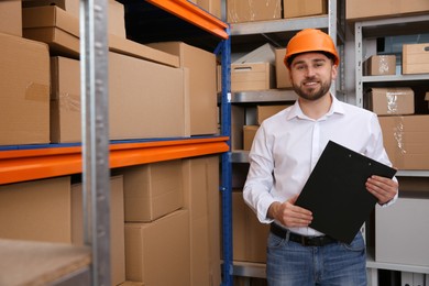 Photo of Young man with clipboard near rack of cardboard boxes at warehouse