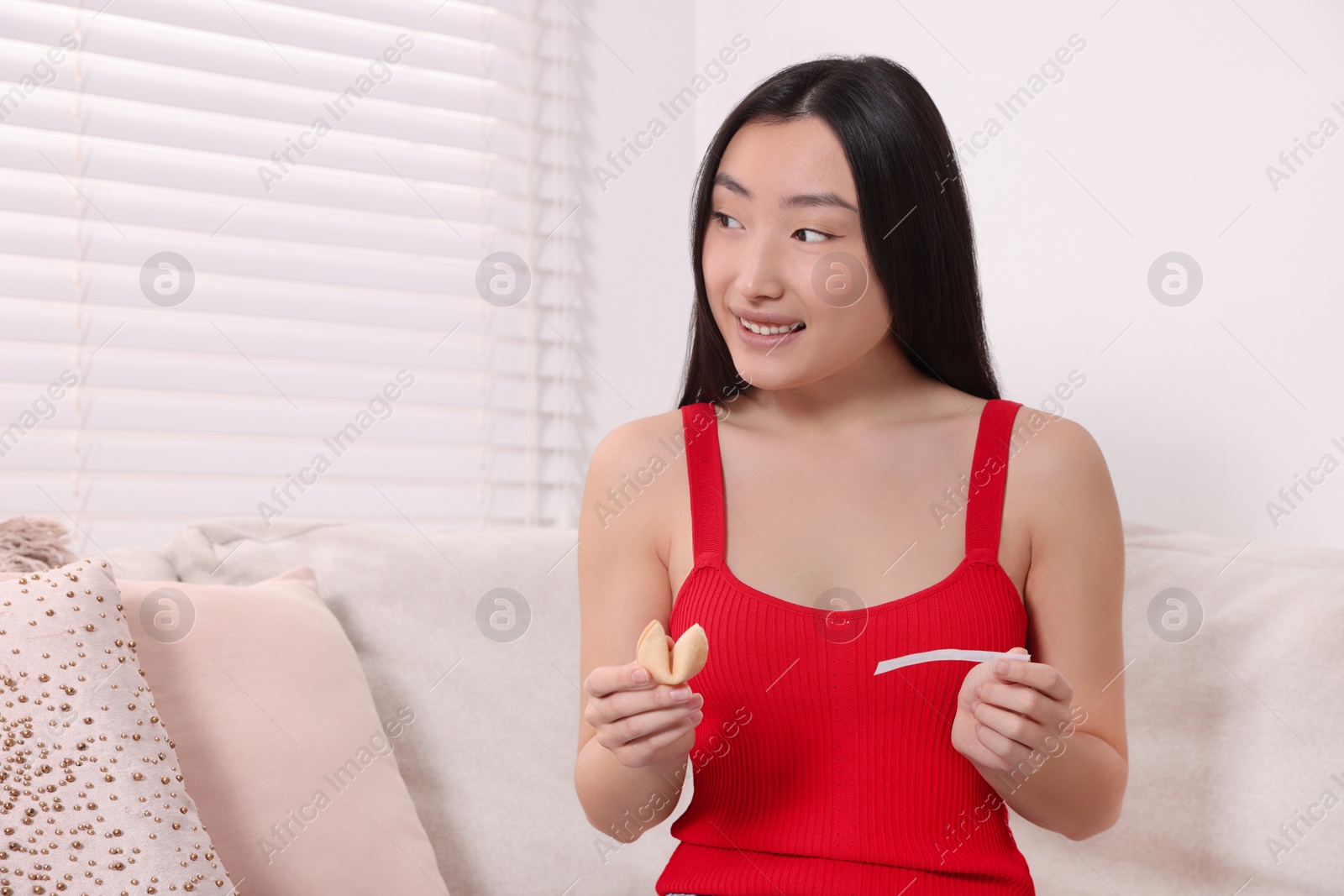 Photo of Asian woman holding tasty fortune cookie with prediction on sofa at home, space for text