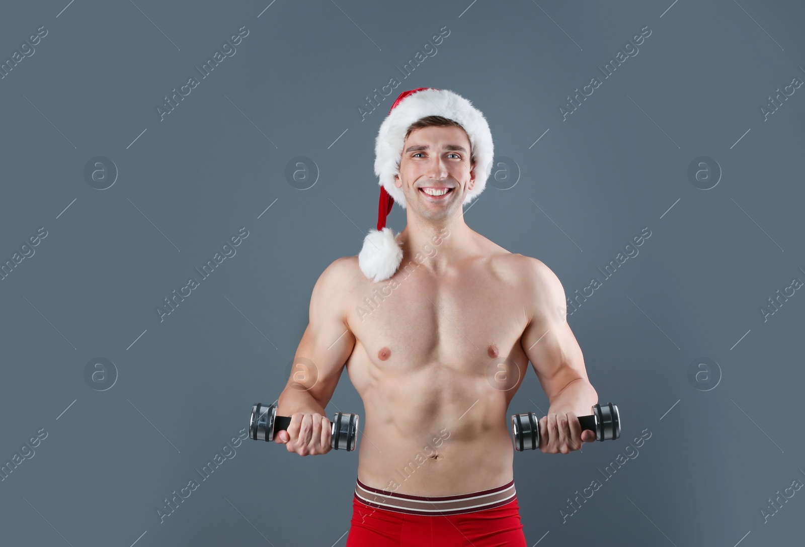 Photo of Young muscular man in Santa hat with dumbbells on gray background