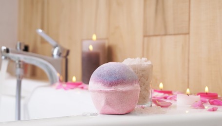 Photo of Colorful bath bomb, sea salt, flower petals and burning candles on white tub in bathroom