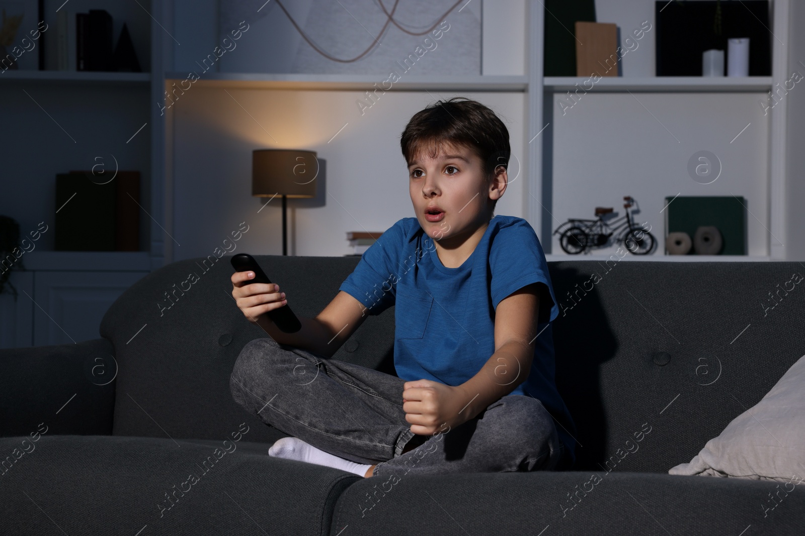 Photo of Emotional boy watching TV and holding remote control on sofa at home