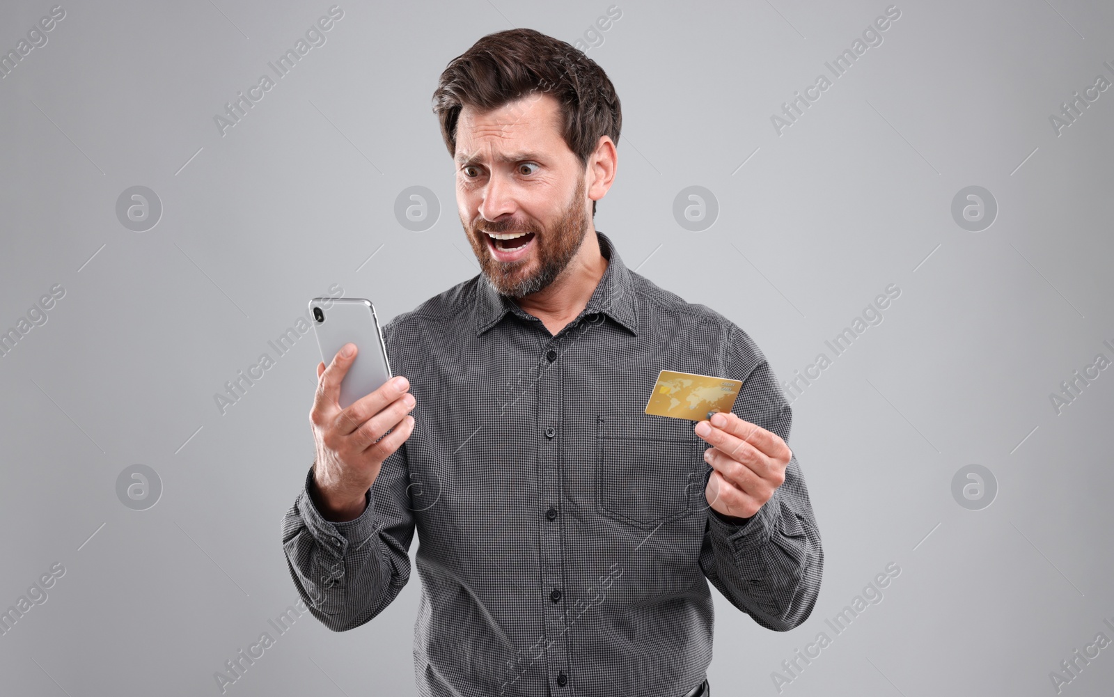 Photo of Emotional man holding smartphone and credit card on light grey background. Be careful - fraud