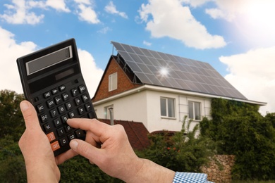 Image of Man using calculator against house with installed solar panels. Renewable energy and money saving