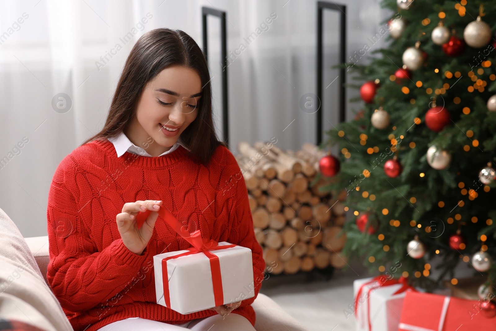 Photo of Smiling woman opening gift near Christmas tree indoors