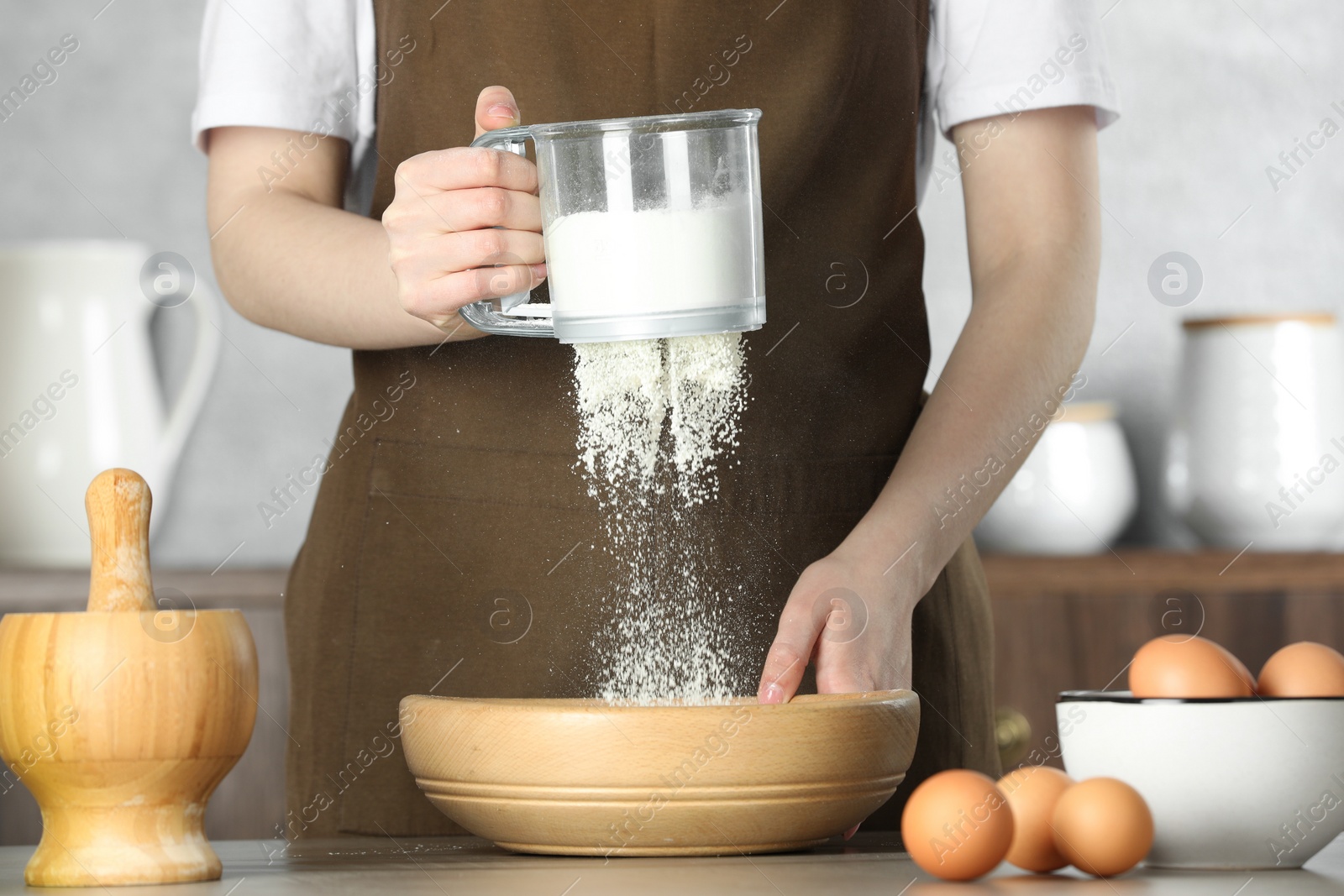 Photo of Woman sieving flour into bowl at table in kitchen, closeup