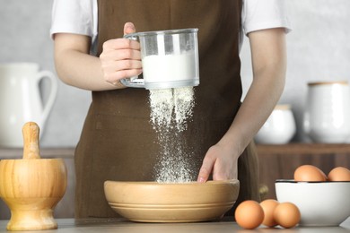 Photo of Woman sieving flour into bowl at table in kitchen, closeup