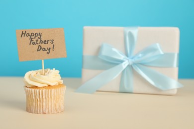 Happy Father's Day. Tasty cupcake with greetings and gift box on beige table, closeup. Space for text