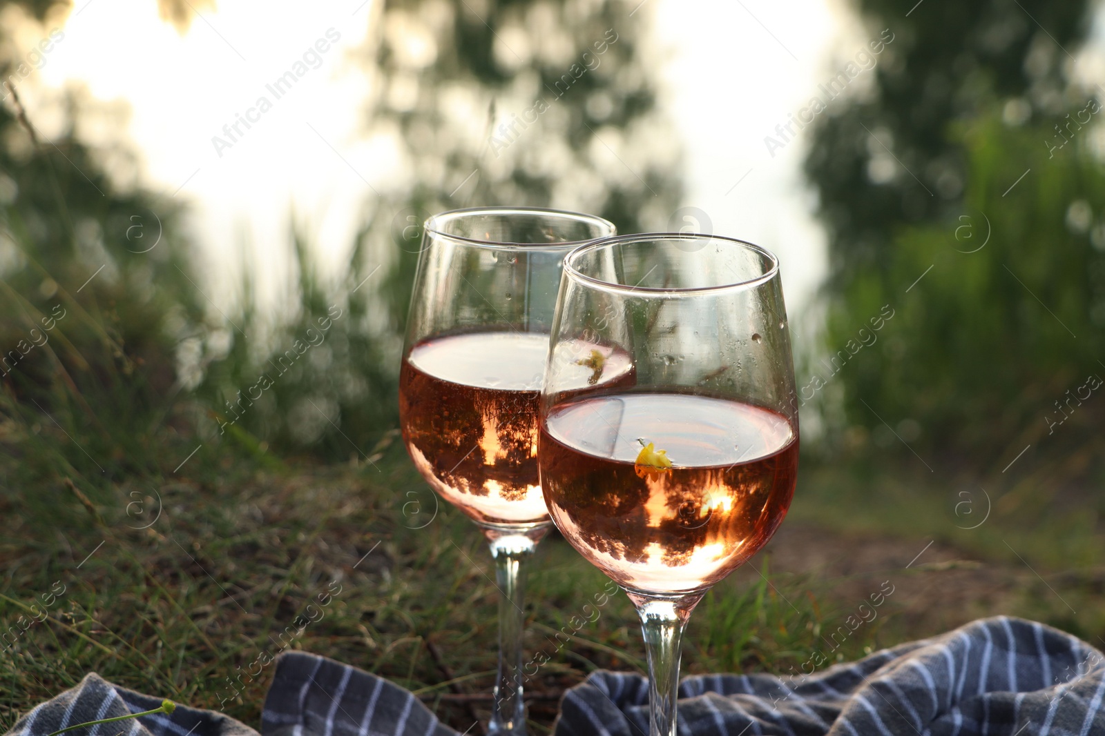 Photo of Glasses of delicious rose wine on picnic blanket near lake, closeup