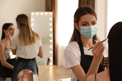 Photo of Makeup artist in protective mask working with woman indoors. Preventive measures during COVID-19 pandemic