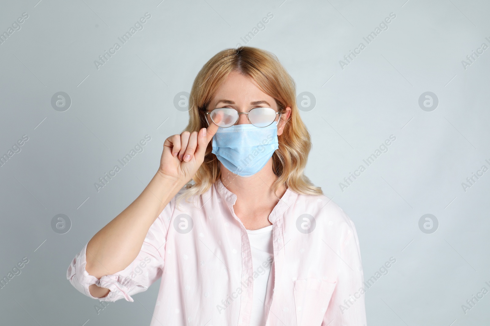 Photo of Woman wiping foggy glasses caused by wearing medical mask on light background