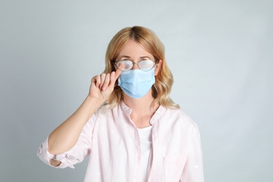 Woman wiping foggy glasses caused by wearing medical mask on light background