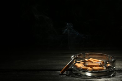 Photo of Glass ashtray with stubs and smoldering cigarette on dark wooden table against black background. Space for text