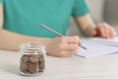 Financial savings. Woman making notes at white wooden table indoors, focus on glass jar with coins