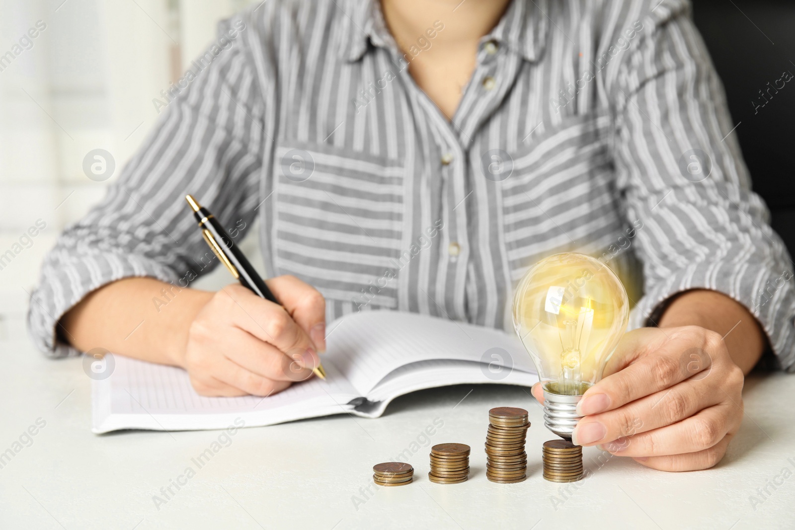 Photo of Woman with light bulb, notebook and coins at white table, closeup. Power saving