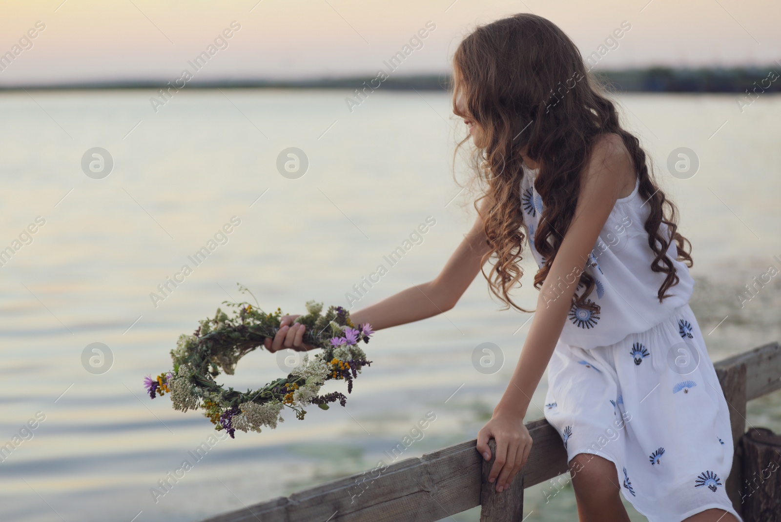 Photo of Cute little girl holding wreath made of beautiful flowers near river in evening