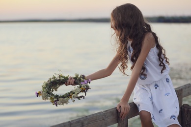 Cute little girl holding wreath made of beautiful flowers near river in evening