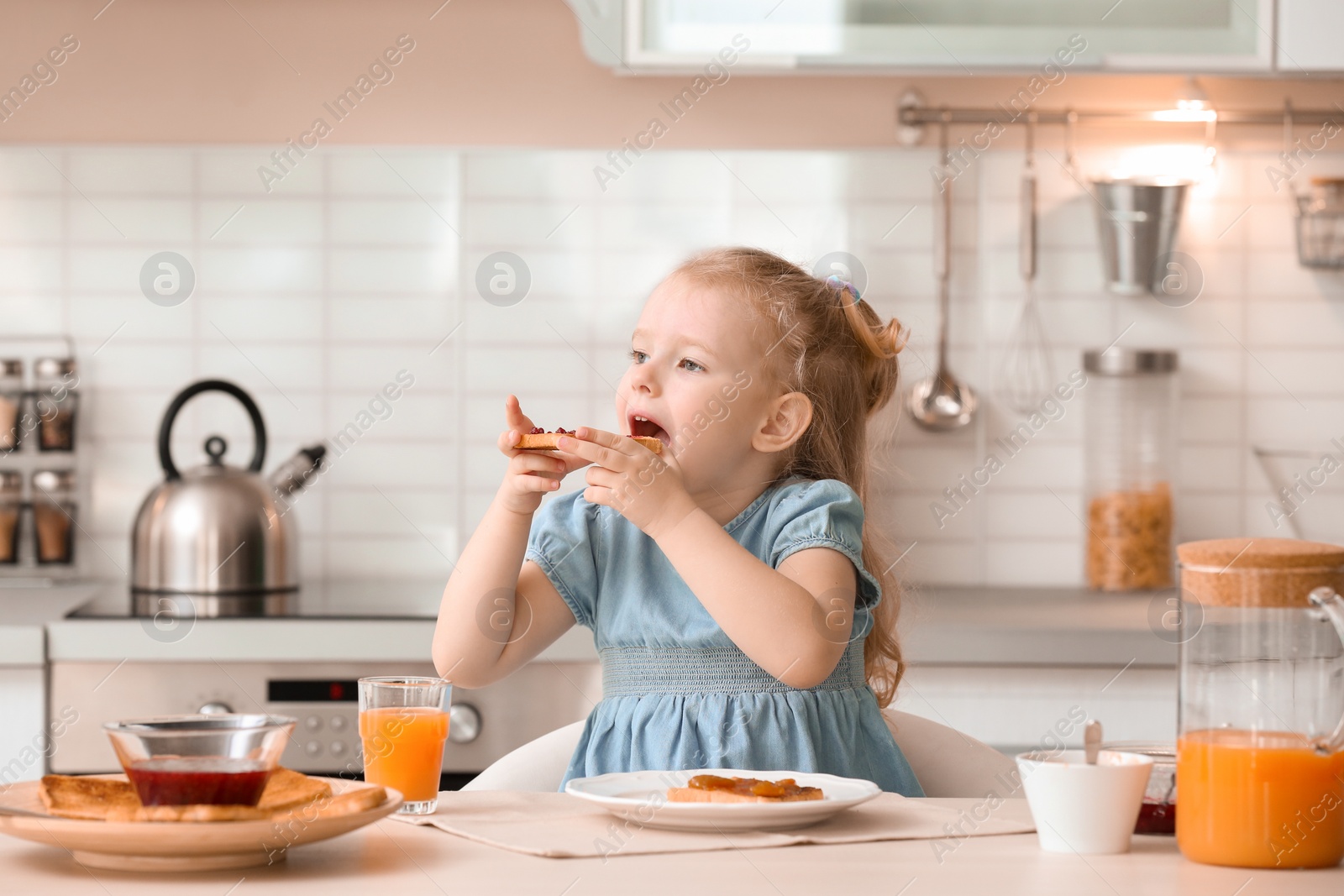 Photo of Adorable little girl eating tasty toasted bread with jam at table in kitchen