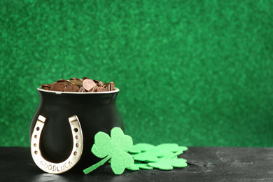 Photo of Pot of gold coins, horseshoe and clover leaves on black stone table against green background, space for text. St. Patrick's Day celebration