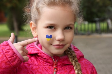 Photo of Little girl with drawing of Ukrainian flag on face in heart shape outdoors
