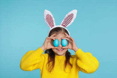 Photo of Little girl in bunny ears headband holding Easter eggs near eyes on color background