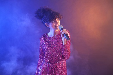 Photo of Beautiful young woman with microphone singing on color background in neon lights and smoke